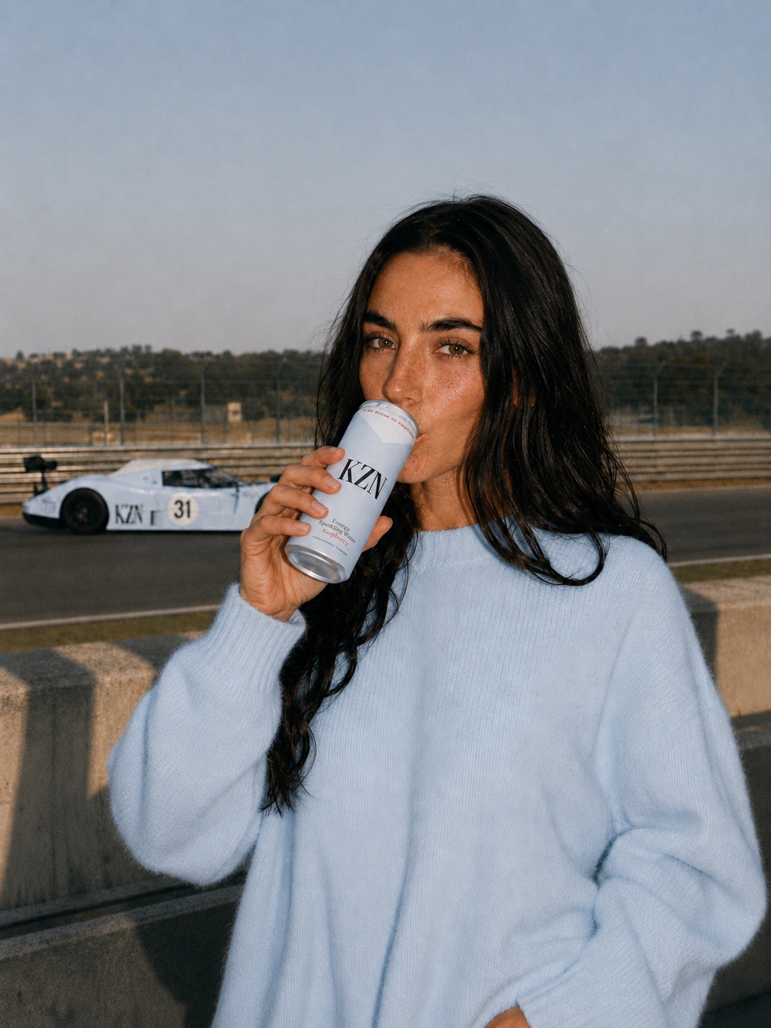 Woman in a pale blue KZN knit drinking KZN Energy beside the KZN race car